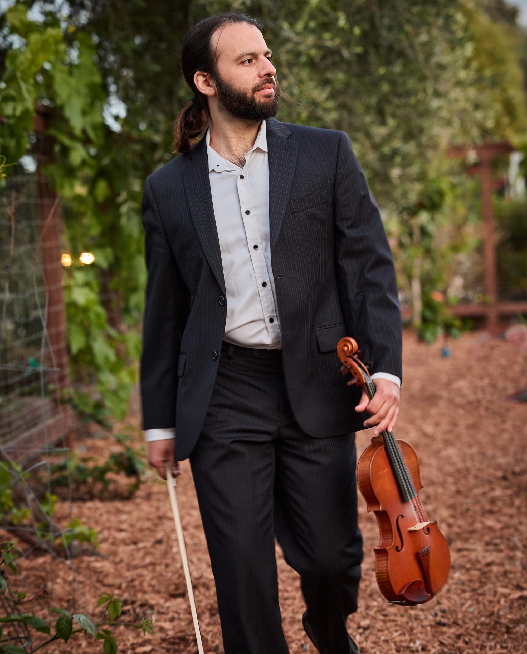 Daniel walking on a tree-lined path in a dark suit, carrying a violin and bow